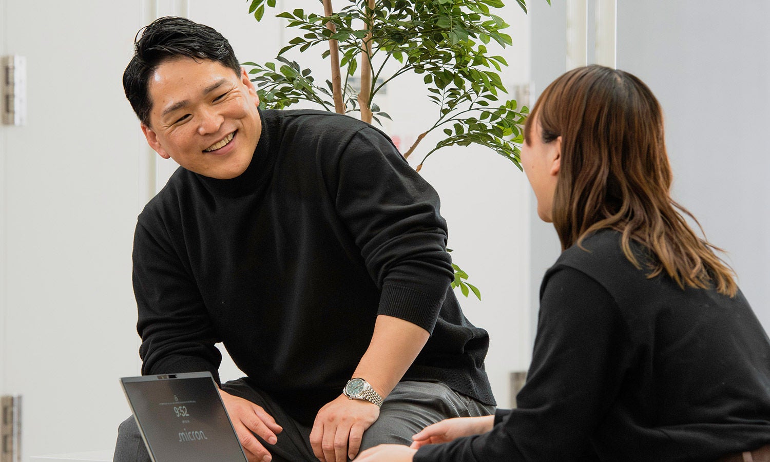 Person leaning forward and talking with a colleague in a casual office setting.