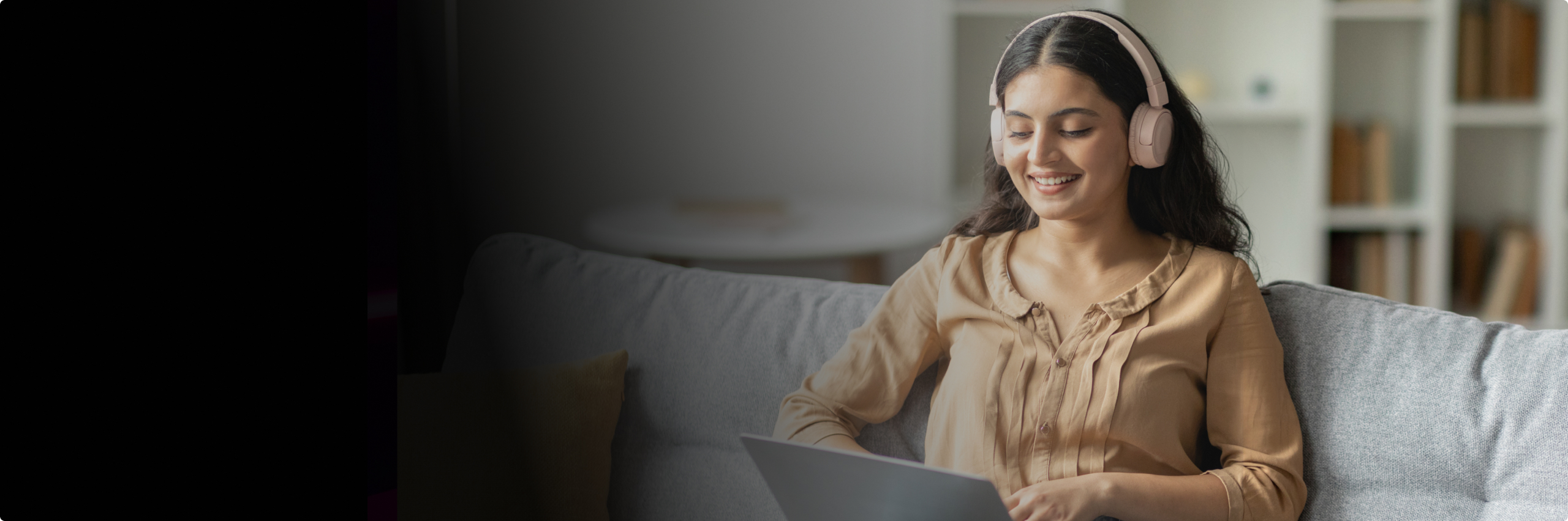 A woman sitting on her couch with headphones and working on a laptop while smiling.