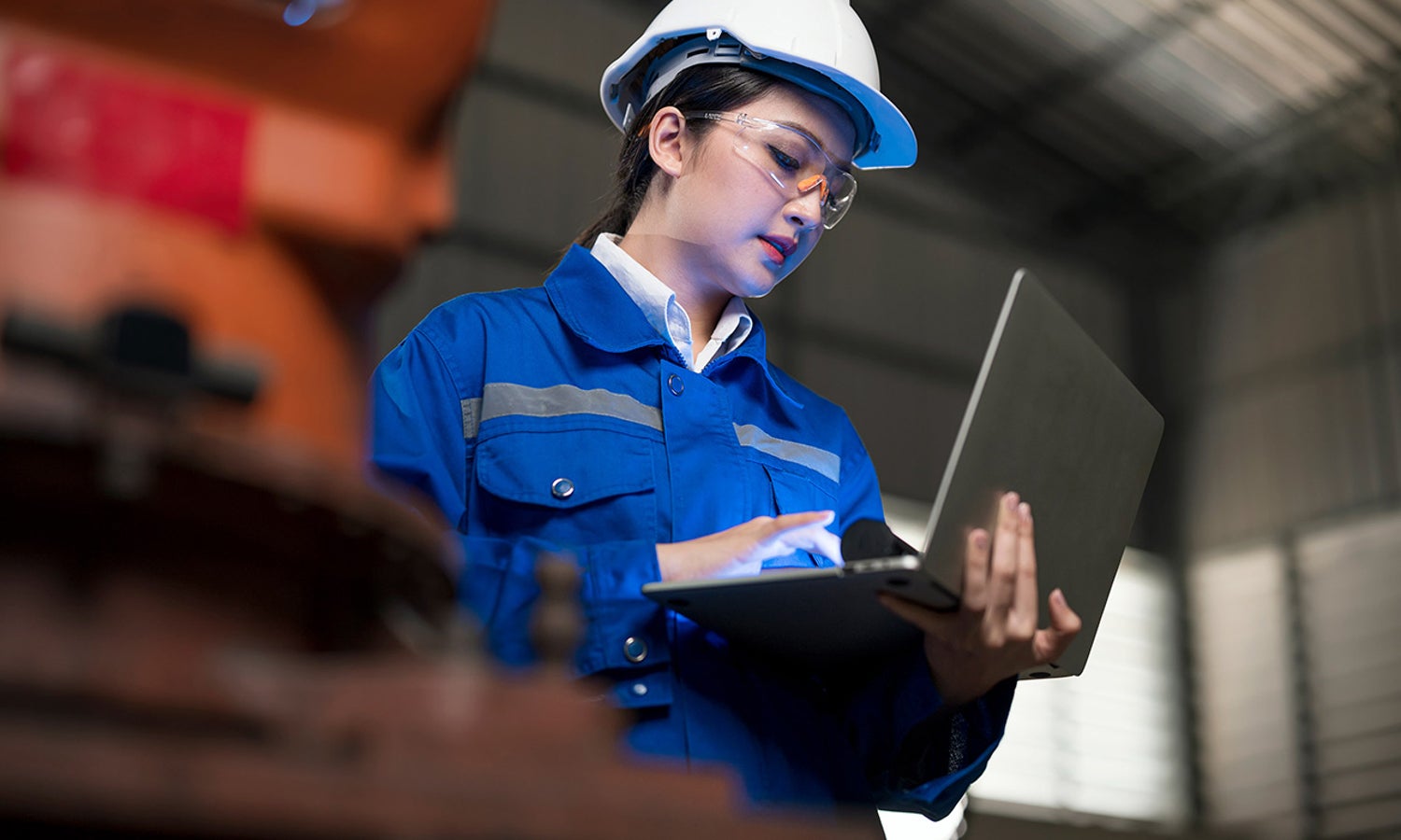 Woman worker, in a hardhad, standing up using her laptop on a manufacturing environment