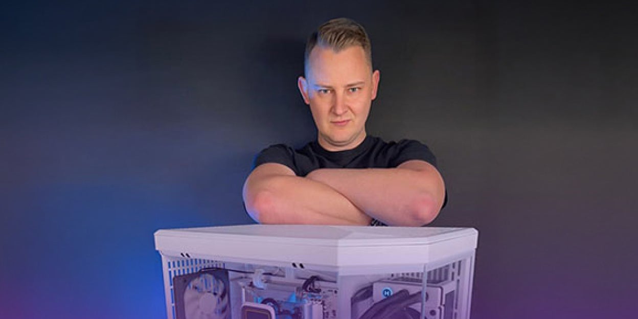 Person standing with arms crossed behind a white custom-built PC featuring visible internal components and RGB lighting, set against a dark background with subtle purple and blue accents.