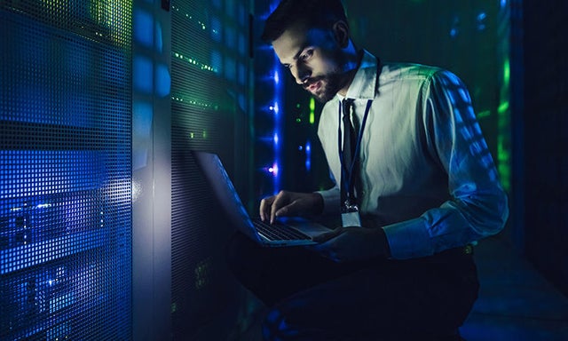 Male worker in dimly lit server room, holding a laptop