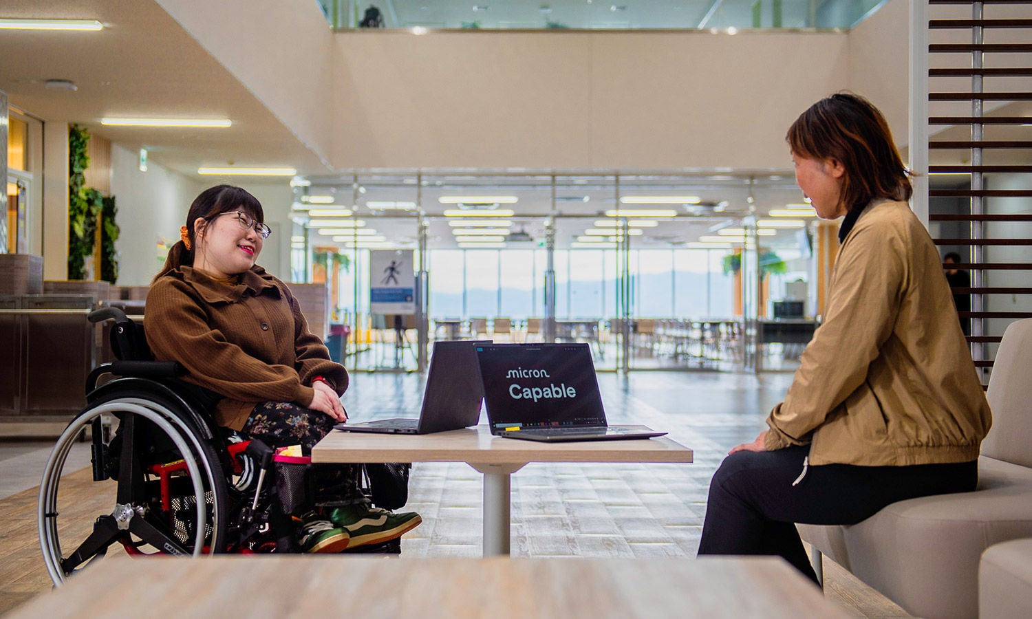 Two people having a conversation beside a laptop in a modern office lobby; one person is seated in a wheelchair.