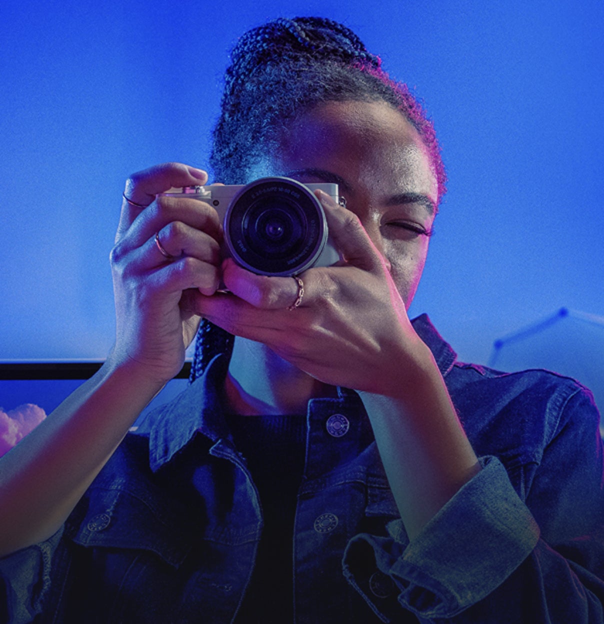 Person wearing a denim jacket is holding a camera up to their face, preparing to take a photo. The background is illuminated with blue and purple lighting, creating a vibrant, studio-like atmosphere.