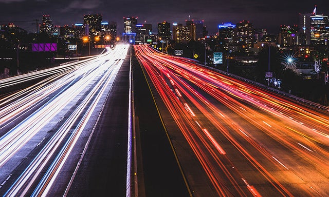 Car headlights and taillights streaking by on a freeway at night with a city in the background