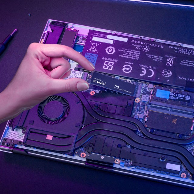 A hand installing a Crucial T710 NVMe M.2 SSD into the motherboard slot on a laptop with the rear panel removed.