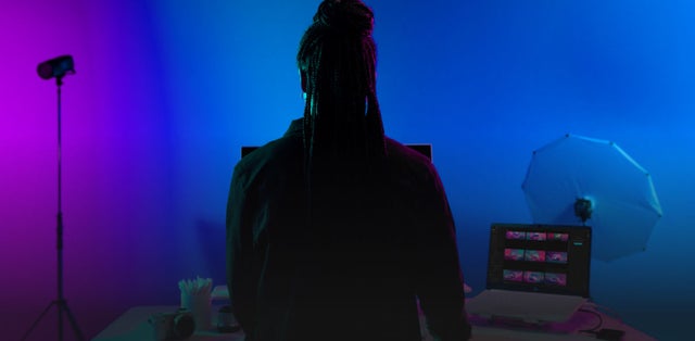 Person with long braided hair is standing in front of a vibrant studio setup featuring purple and blue lighting. A photography light stand is positioned on the left, and a white umbrella reflector is visible on the right, creating a professional photo shoot environment.