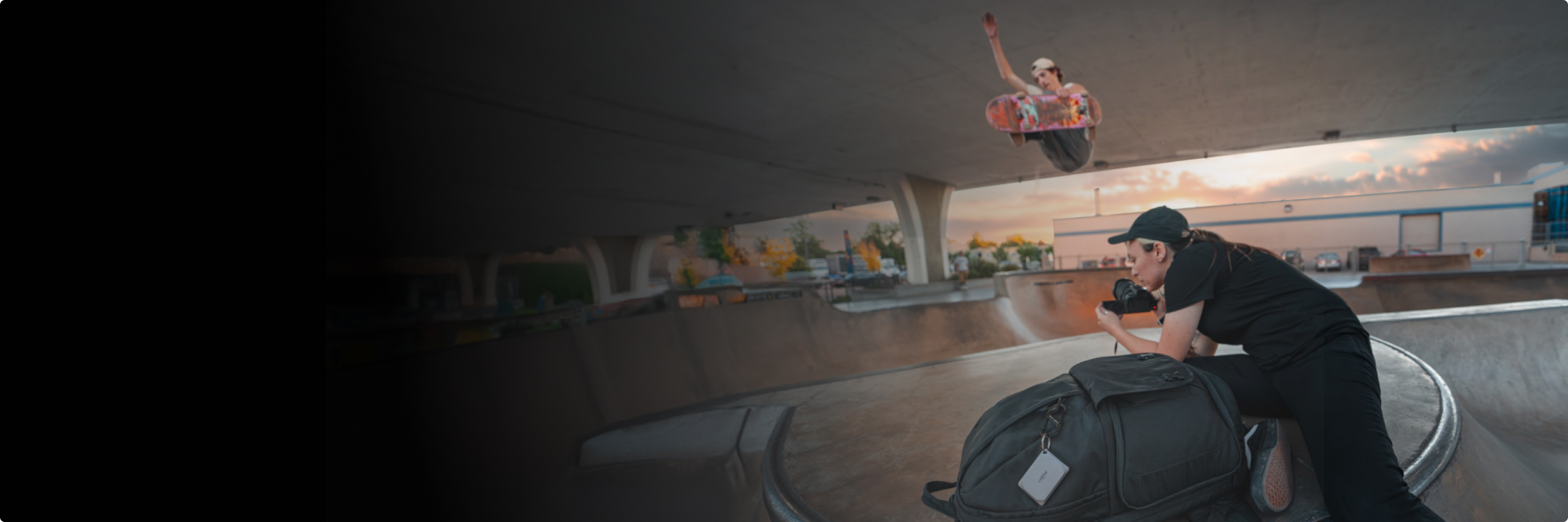A photographer in a leaning/lying position on top an underpass skatepark quarter pipe taking a photo of a skateboarder midair with a sunset in the background.