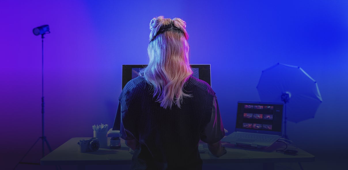 紫と青の照明に照らされたスタジオの前に立つ、長い編んだ髪の人物。左側に置かれた撮影用ライトスタンド、右側に見える白い傘型リフレクターが、プロの撮影現場の雰囲気を演出している。