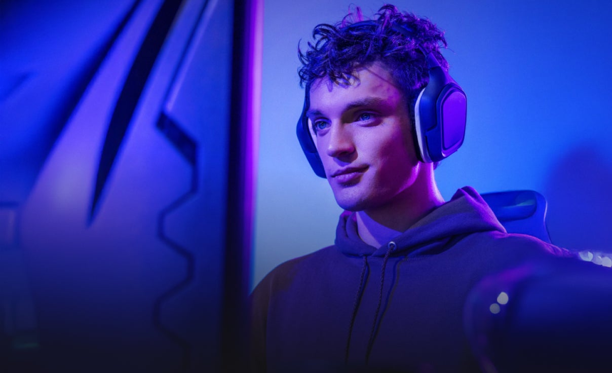 Person wearing large over-ear gaming headphones and a hoodie, seated in front of a monitor in a room illuminated with vibrant blue and purple lighting, creating a modern gaming setup atmosphere.