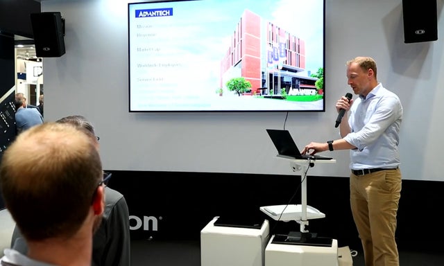 Man speaking into a microphone looking at a laptop with a large monitor in the background