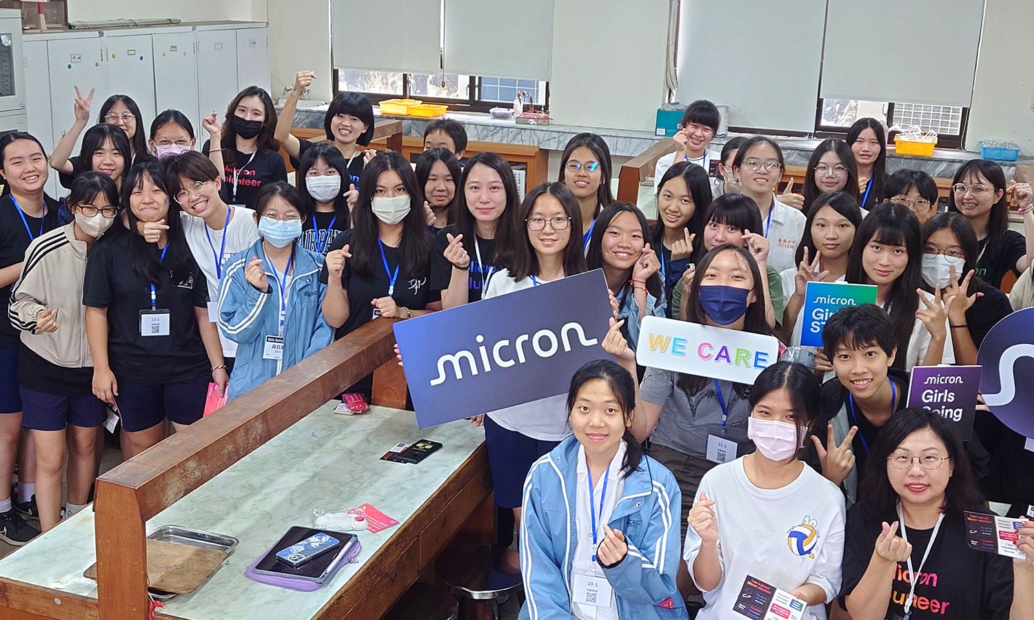 Students participating in a Micron Girls in STEM event, holding Micron and We Care signs in a classroom.