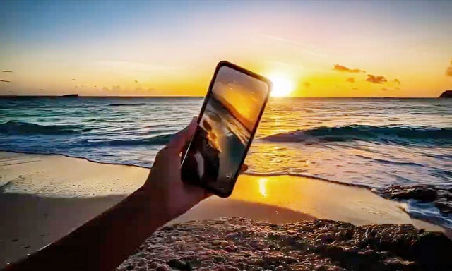 Hand holding a smartphone displaying a beach sunset over ocean waves