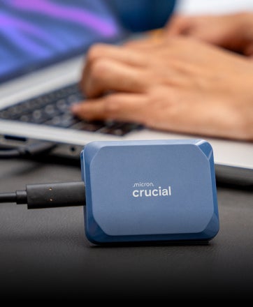  Una SSD portátil Crucial X9 Pro de 4 TB colocada verticalmente en un escritorio frente a un ordenador portátil.