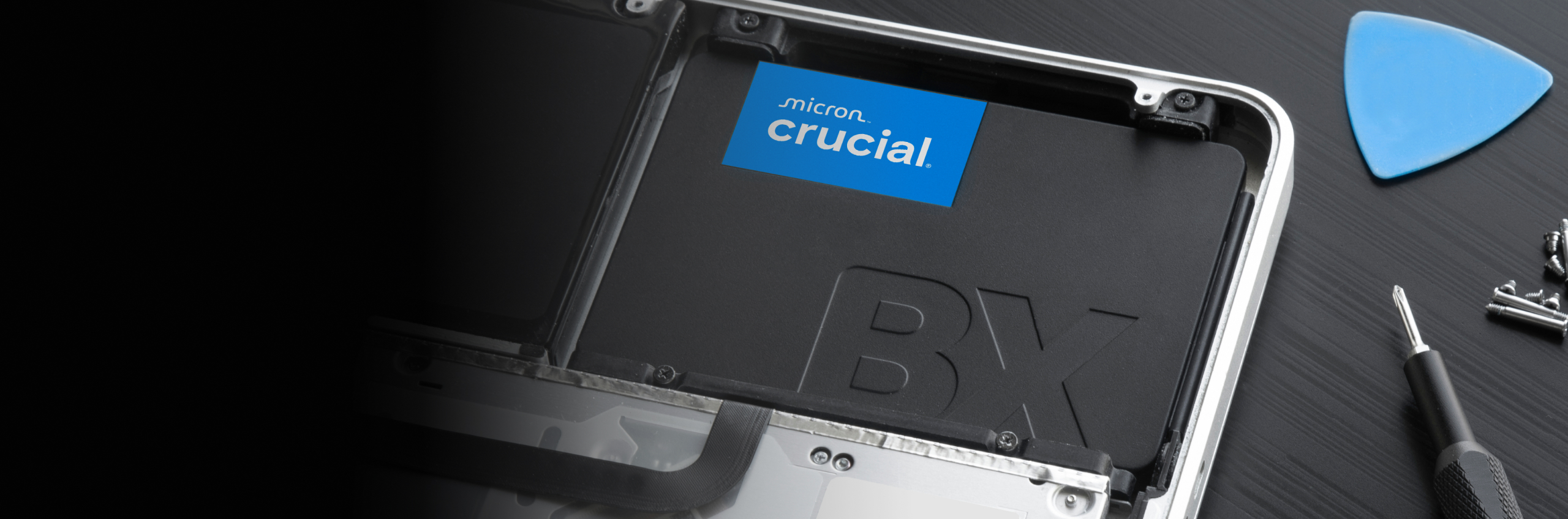 Close shot of the corner of an opened laptop next to a screwdriver and small, removed screws. A Crucial 2.5-inch SATA SSD is installed in the laptop.