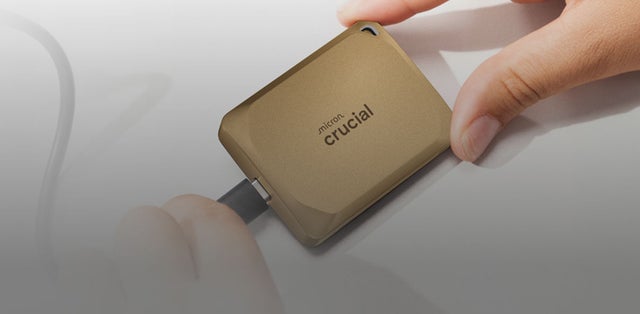 Close-up of a gold Crucial X10 Pro portable SSD connected via a cable, with a hand holding the device against a clean white background.