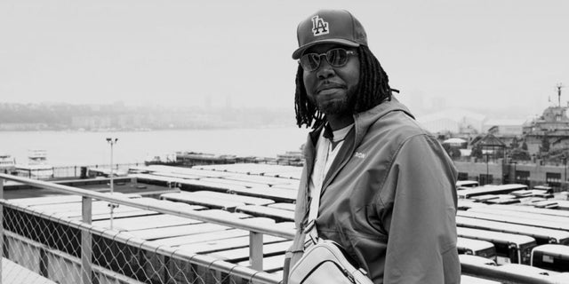 Person wearing a baseball cap and a jacket, standing near a metal railing overlooking a waterfront with rows of docked boats and industrial structures in the background. The man has a crossbody bag and long braided hair.
