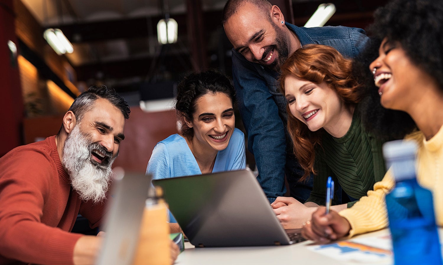 A group of diverse, smiling and laughing people, sitting around a laptop.