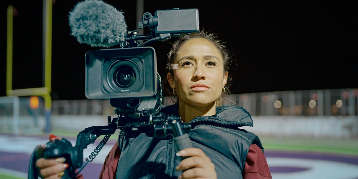 Sarina Soriano standing on a football field and staring into the distance while holding a professional video camera.