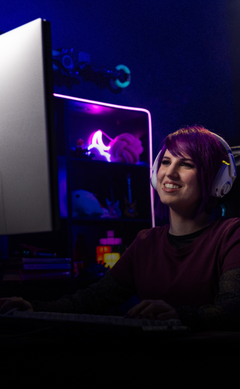  A smiling woman with headphones sitting and playing on a gaming PC.